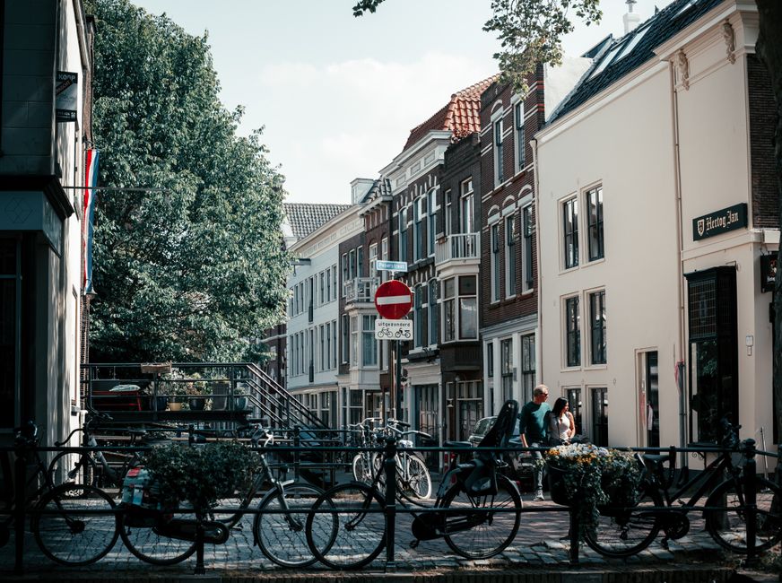 This image depicts a European urban street scene with a row of multi-story residential buildings featuring a mix of brick and stucco facades, some with decorative window trims and balconies. The buildings vary in height and color, including white, dark brown, and red brick finishes. In the foreground, a black metal bike rack is filled with bicycles parked along the sidewalk. Two pedestrians are visible walking and engaging near the curb; a man and a woman, casually dressed, with the woman holding a bag. To the right, a small tree with dense foliage partially shades the scene, and there are planter boxes with flowers attached to the bike rack. A street sign indicating a no-entry zone for vehicles is mounted on a pole, with additional smaller signs below it, including bicycle route markers. The environment suggests a quiet, well-maintained neighbourhood with natural light illuminating the scene, appropriate for a context of private or independent waste collection services like those offered by House Clearance Holland Park, especially for complex or on-site rubbish removal tasks in residential areas.
