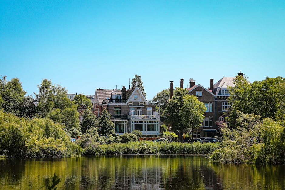 A daytime scene showcasing a collection of residential houses situated along a riverbank, with several multi-storey buildings featuring traditional brick facades, large sash windows, and pitched tiled roofs. Some houses have prominent chimneys and decorative gables, while others display balconies or terraces facing the water. The foreground includes a calm, reflective body of water bordered by lush green bushes and mature trees, casting gentle shadows on the bank. The area appears well-maintained, with neatly trimmed vegetation and a clear, bright blue sky overhead. This urban riverside setting exemplifies a peaceful neighborhood, where private property and natural environments coexist, providing an ideal backdrop for independent rubbish clearance services by companies such as House Clearance Holland Park, who may handle waste removal for estate cleanups or property upgrades in the area.