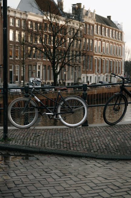 The image depicts a black bicycle with a brown leather saddle and silver accents, securely locked to a black metal railing along a brick-paved pathway. In the background, there is a canal with calm water reflecting the overcast sky and the brick facades of historic townhouses, characteristic of a traditional urban setting. The townhouses are multi-storey, constructed from reddish-brown brick with white window trims, some with decorative cornices and large sash windows, and a few trees with bare branches line the street, indicating a colder season. Adjacent to the railing, a second bicycle with a dark frame and minimal features is also locked in place, indicating a shared practice of on-site bicycle parking in the area. The backdrop includes a bridge over the canal and distant rooftops, contributing to the scene's typical cityscape ambiance. The setting appears quiet and well-maintained, fitting within a neighbourhood where private arrangements for transport security, such as bicycle locks, are common alongside public pathways, and the environment subtly aligns with neighbourhood waste management and clearance activities possible with local or independent collection services like House Clearance Holland Park.