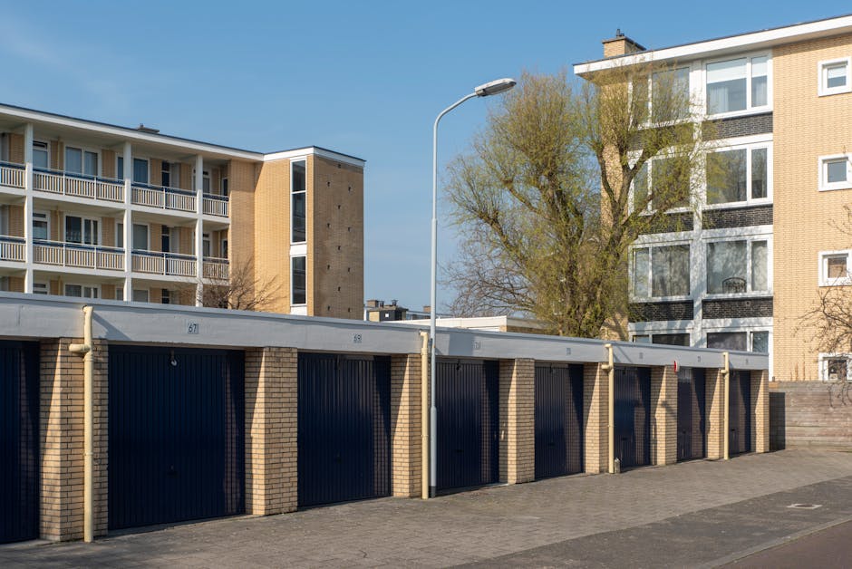 A daytime scene showcasing a collection of residential houses situated along a riverbank, with several multi-storey buildings featuring traditional brick facades, large sash windows, and pitched tiled roofs. Some houses have prominent chimneys and decorative gables, while others display balconies or terraces facing the water. The foreground includes a calm, reflective body of water bordered by lush green bushes and mature trees, casting gentle shadows on the bank. The area appears well-maintained, with neatly trimmed vegetation and a clear, bright blue sky overhead. This urban riverside setting exemplifies a peaceful neighborhood, where private property and natural environments coexist, providing an ideal backdrop for independent rubbish clearance services by companies such as House Clearance Holland Park, who may handle waste removal for estate cleanups or property upgrades in the area.