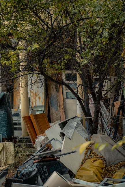 A pile of discarded household items and debris situated outdoors on a patch of ground covered with leaves, with a backdrop of a wooden fence, trees, and an aged building. The waste includes a white deconstructed cabinet or appliance, a yellow bag, wooden panels with a brown finish, and various smaller objects and tools. The environment appears to be a cluttered backyard or garden area adjacent to a residential property, with natural light filtering through the trees. The scene illustrates a typical example of excess rubbish that may be removed via private rubbish clearance services, such as those provided by House Clearance Holland Park, especially for property cleanouts or bulk waste disposal. The arrangement and assortment of materials highlight the need for professional waste handling to manage bulky household refuse or renovation debris in the W11 postcode area.