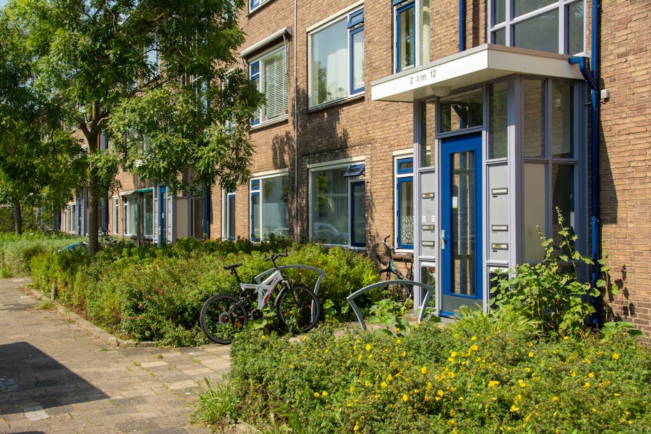 The image shows the entrance to a residential apartment building with a small, modern canopy over a blue-framed glass door. To the right of the door, there are four metallic mailboxes mounted on the brick wall, and the canopy features white panels displaying the number '2' and the note 'tm 12'. Surrounding the entrance, there is a neatly maintained green shrubbery bed with small yellow flowers, framed by a stone-paved pathway. Several bicycles, including a mountain bike with a white frame and an adjacent bike with a black frame, are parked near the garden area, leaning against a curved metal bike rack. In the background, there are large windows with white framing, some partially open, and the building's exterior wall is constructed from reddish-brown bricks. A tall deciduous tree on the left provides partial shade, casting shadows onto the sidewalk and greenery, and the scene depicts a calm, urban residential environment that may be associated with private waste management or alternative rubbish collection solutions. The image is well-lit by natural daylight, emphasizing the textures and colors of the materials and surroundings.