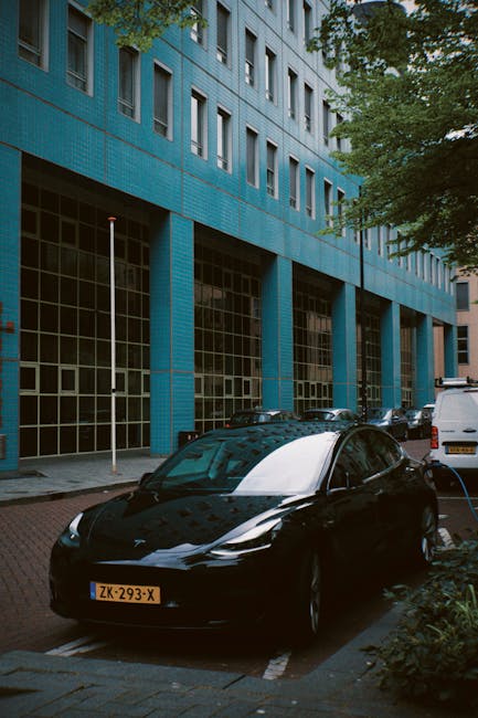 A black electric car with a sleek, glossy finish is parked on a city street in front of a modern blue building with large glass windows on the ground floor. The car, situated in a designated parking space, has a yellow license plate and appears to be charging, with a charging cable connected to its side. The building's exterior features a grid of small square tiles painted in a vibrant blue color, with multiple Windows above. To the right, a white van is parked parallel to the electric vehicle. Surrounding the scene are lush green trees providing partial shade, with branches extending over the parked vehicles. The street surface is paved and lined with a white dashed line marking parking boundaries. The natural light suggests daytime, and the overall scene reflects an urban environment where private, alternative waste or vehicle-related services such as electric vehicle charging are common, aligning with the context of independent collection and on-site management that House Clearance Holland Park might support.